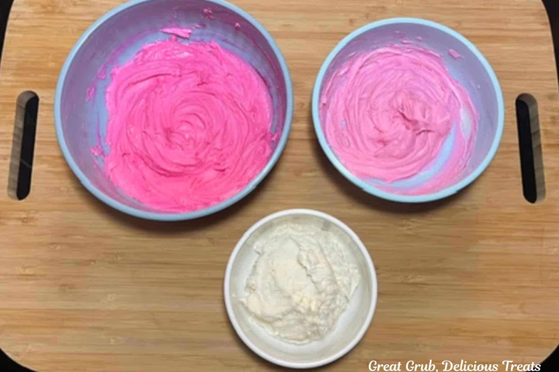 A wood surface with three bowls filled with buttercream frosting, one white, one light pink, and one bright pink.