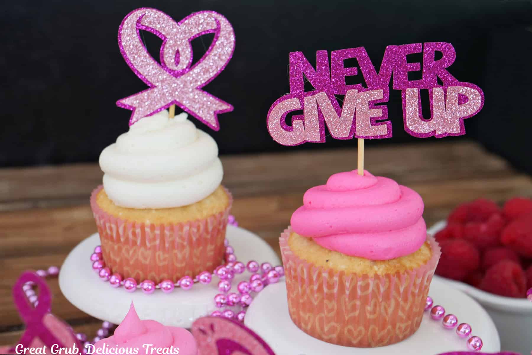 Two small plates with one cupcake on each, one with pink frosting and one with white frosting, with breast cancer awareness toppers, one heart, and one that say "never give up."