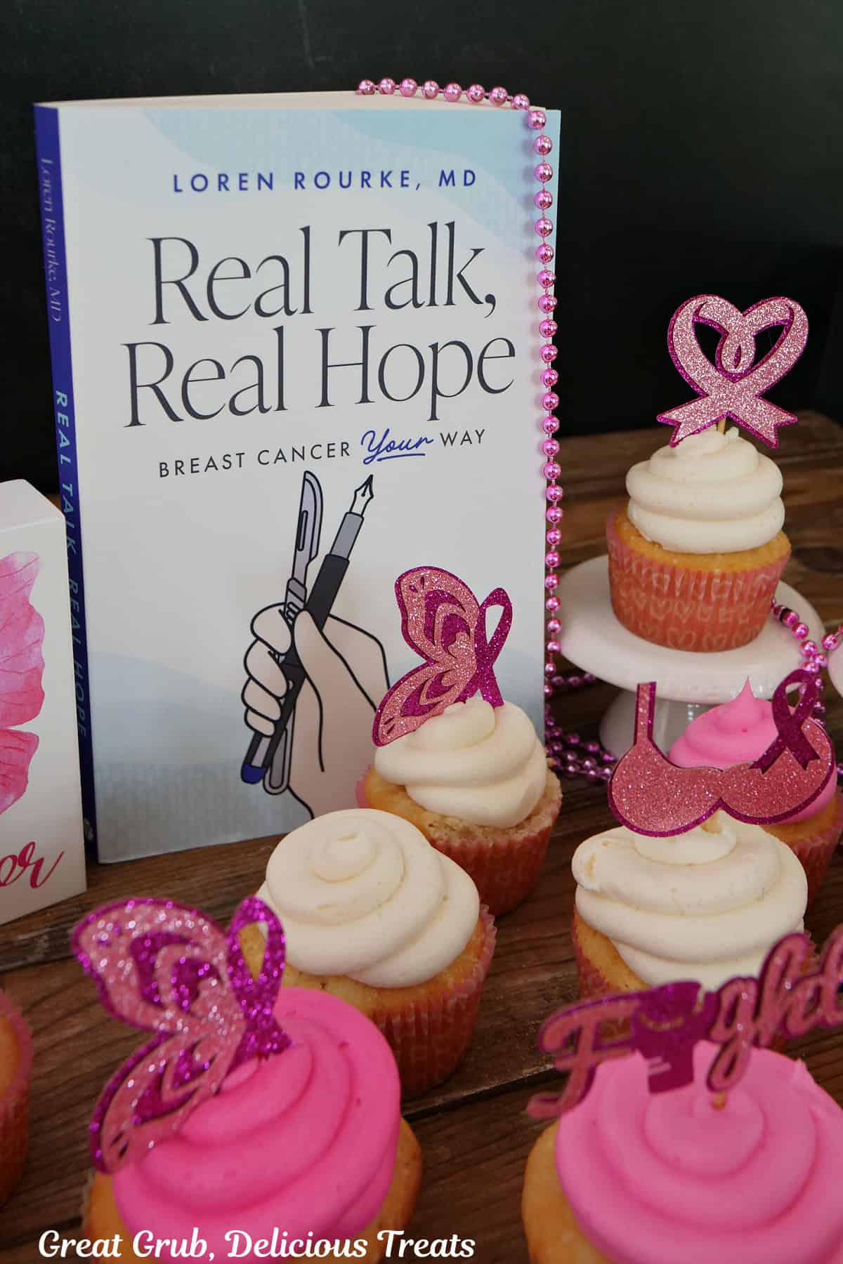 White cupcakes with pink and white frosting with breast cancer awareness toppers, and Dr. Loren Rourke's Real Talk, Real Hope book in the background.