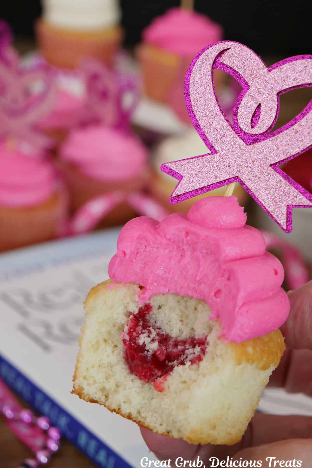 A white cupcake with pink buttercream frosting with a bite taken out showing the raspberry filling, and it has a heart paper topper on top with more cupcakes in the background.