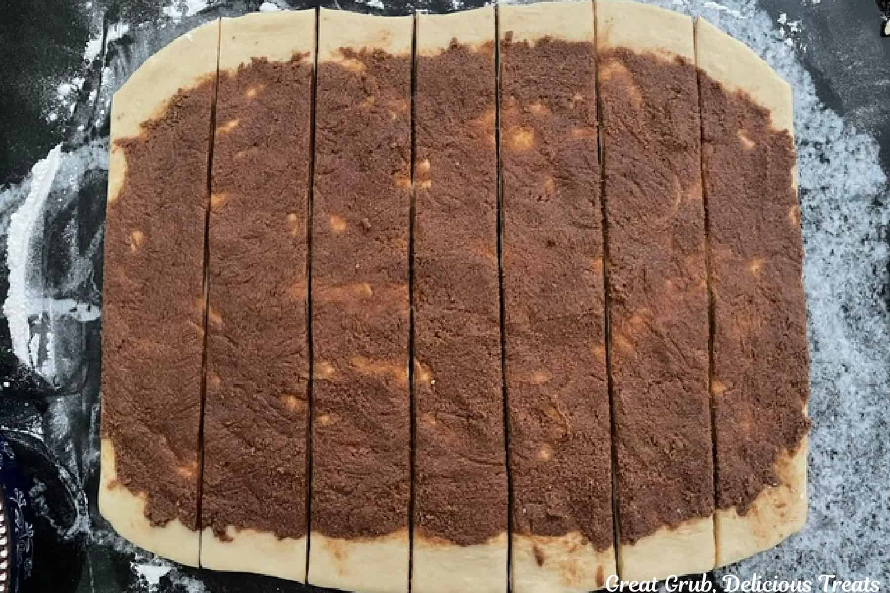 A black countertop with cinnamon roll dough rolled out topped with cinnamon filling, and cut into seven strips.