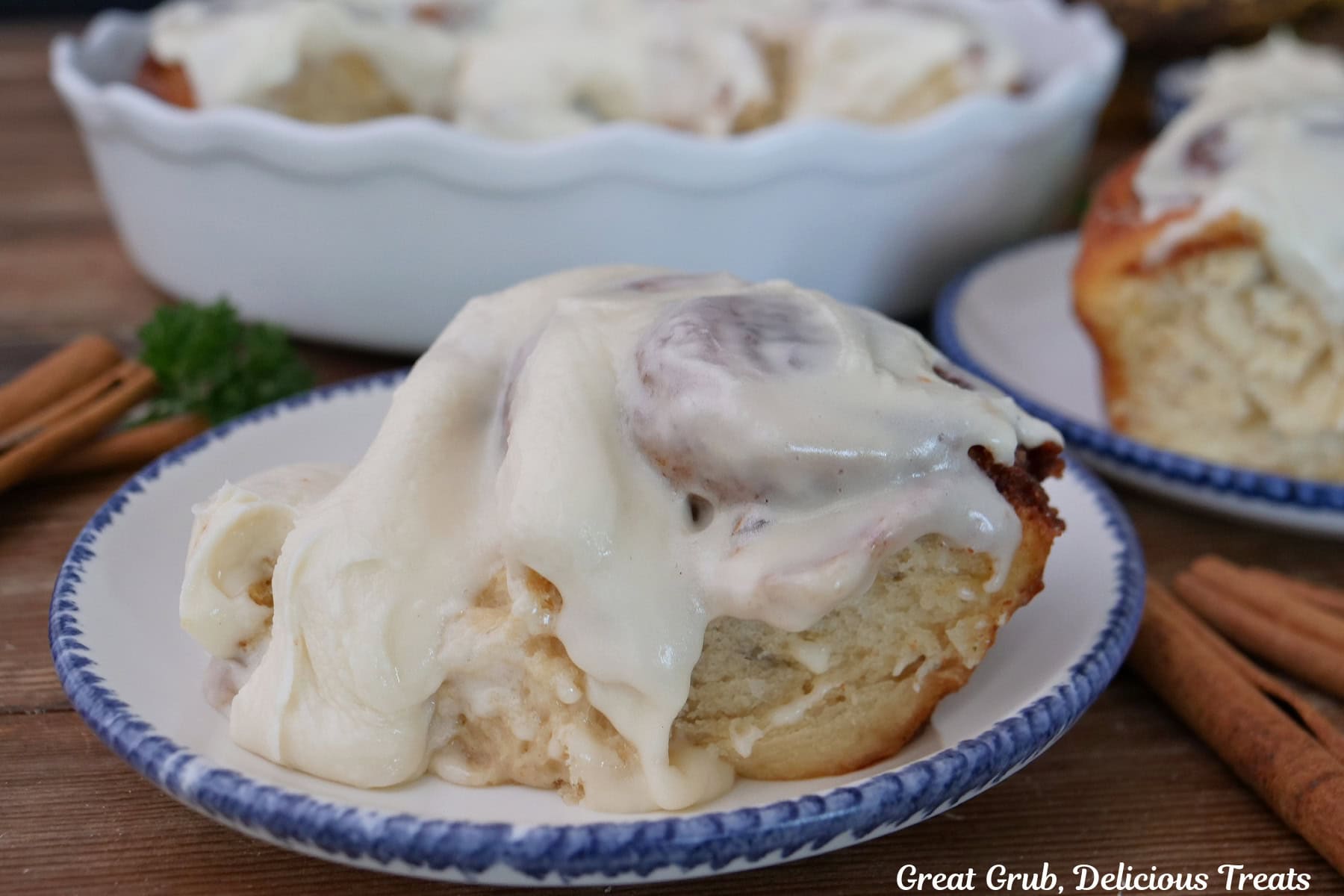 A white plate with blue trim with a banana cinnamon roll on it topped with cream cheese icing, with more cinnamon rolls in the background.