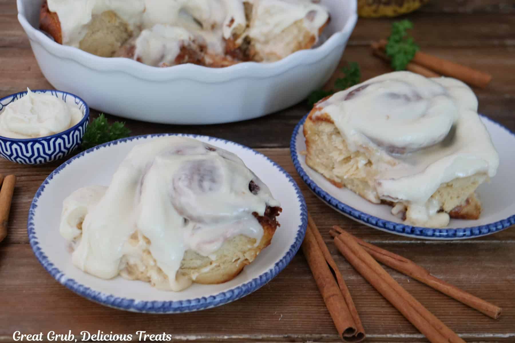 A wood surface with two white plates with blue trim with a cream cheese frosted banana cinnamon roll on them, with more cinnamon rolls in the background.