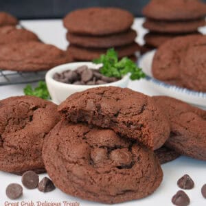 A white surface with dark chocolate cookies randomly placed about with a small white bowl filled with baking chips.