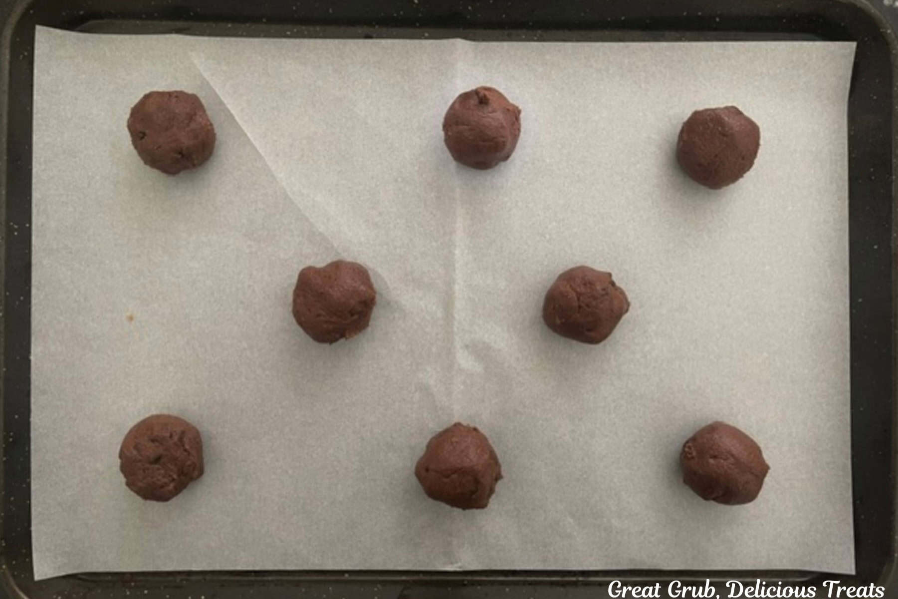 A baking sheet lined with parchment paper with eight chocolate cookie dough balls on it.