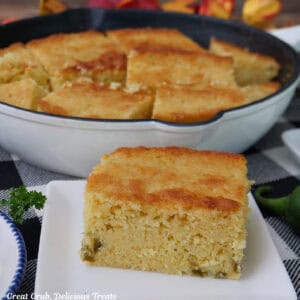 A white square plate with a serving of jalapeno cornbread on it with a cast iron pan skillet filled with cornbread in the background.