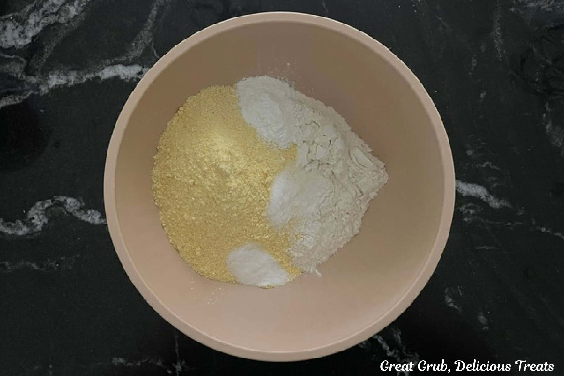 A black countertop with a tan mixing bowl with the dry cornbread ingredients in it.