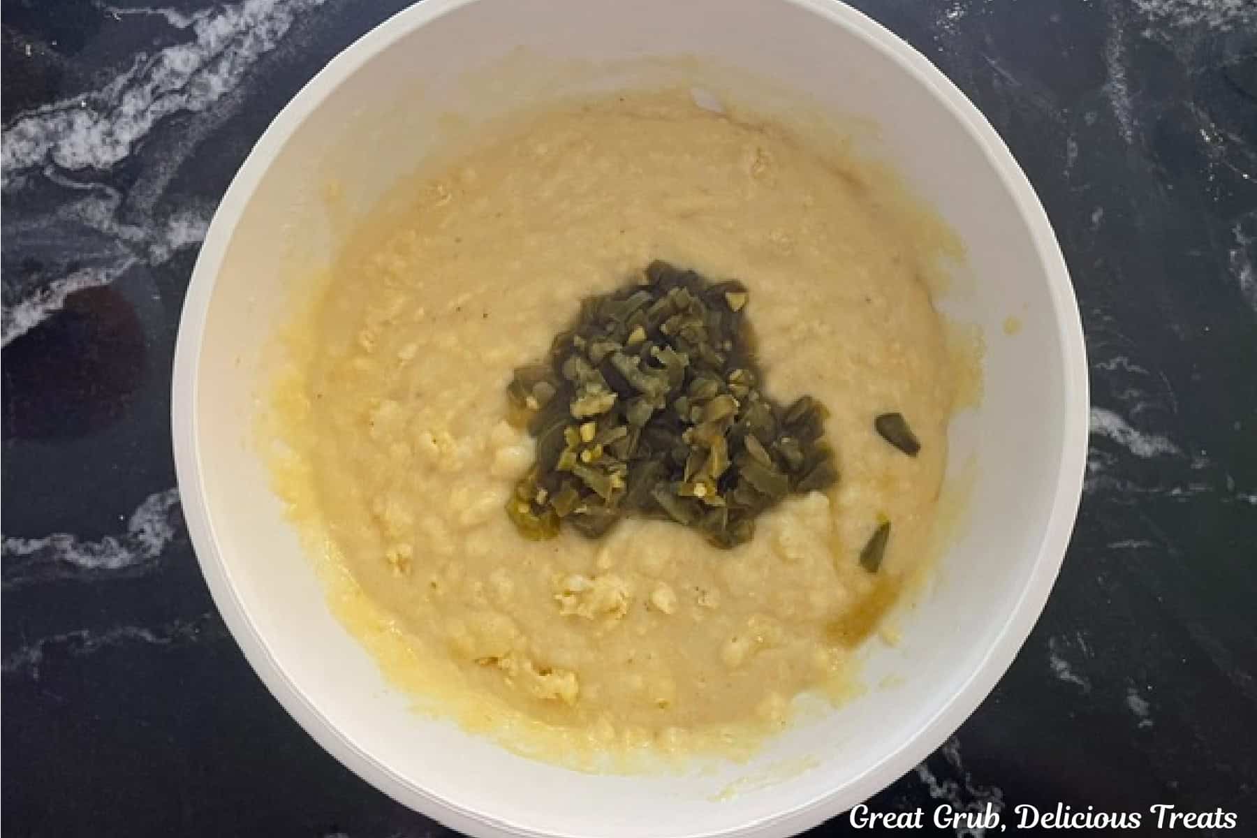 A black countertop with a white mixing bowl with diced jalapenos added to the cornbread batter.