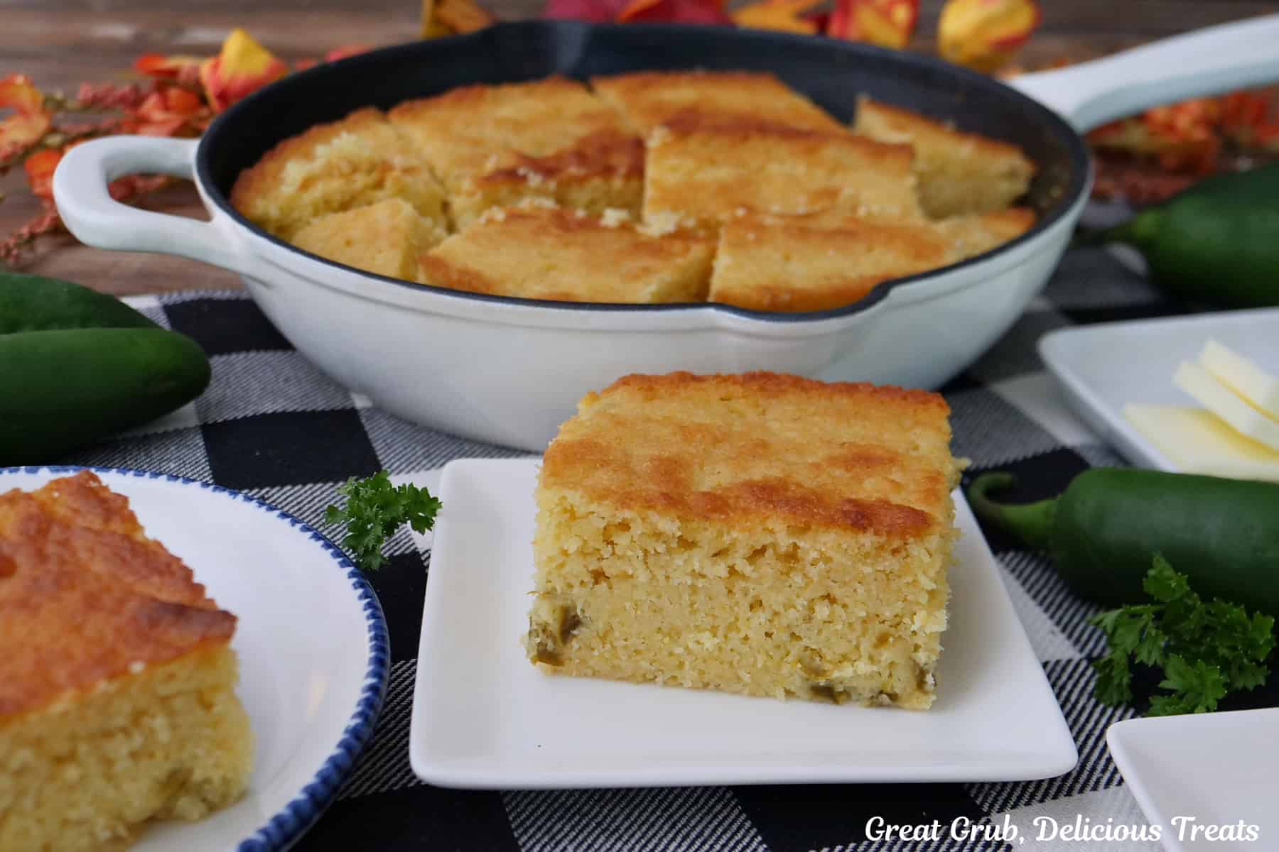 Two white plates with a serving of cornbread on them, and a white cast iron skillet in the background filled with jalapeno cornbread.