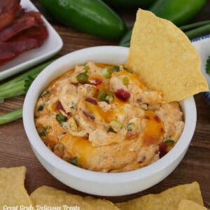 A white bowl filled with jalapeno popper dip with a tortilla chip in the right side of the dip with more chips in the forefront and bacon and jalapenos in the background.