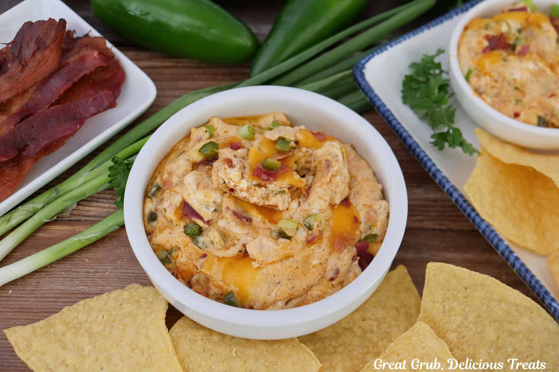 A white bowl filled with a serving of jalapeno popper dip with bacon, jalapenos, green onions, and tortilla chips in the photo.