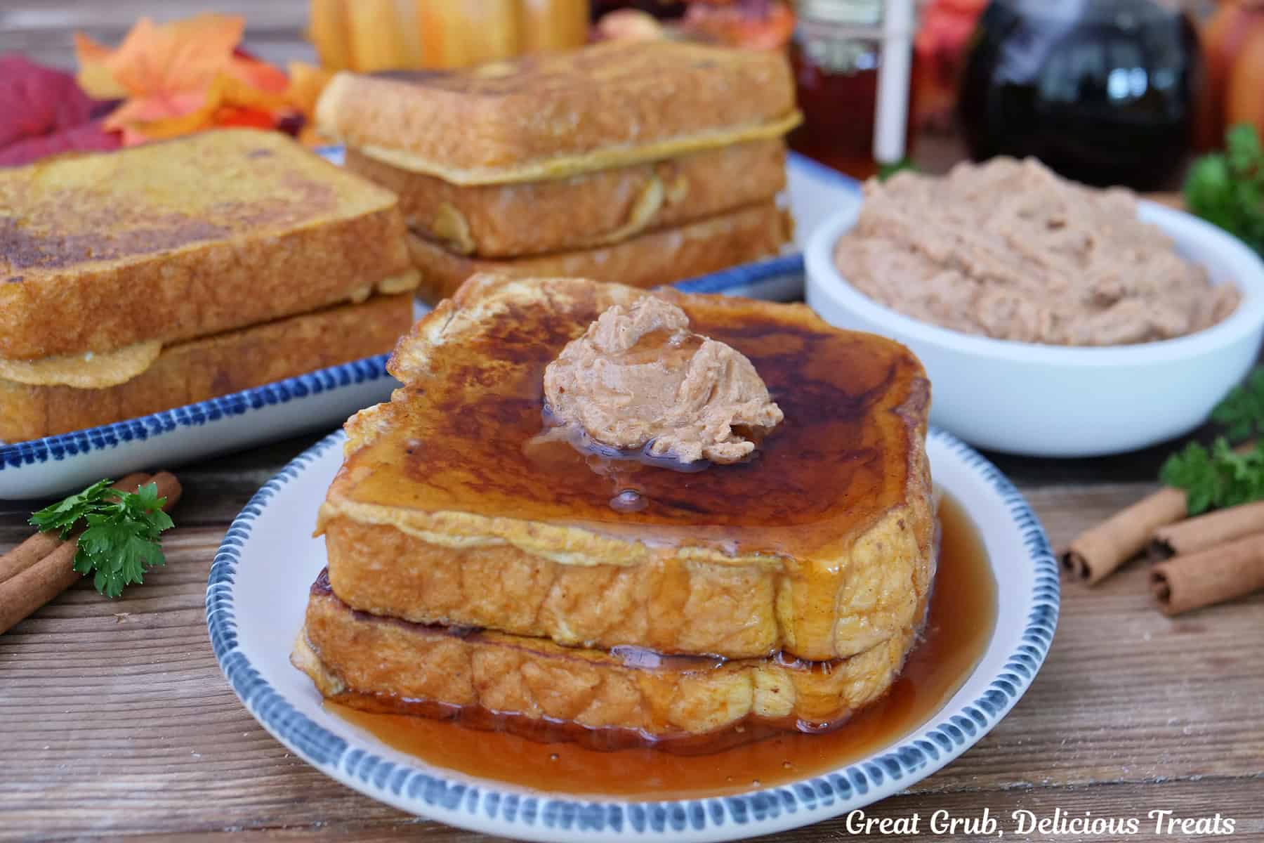 A wood surface with a blue trimmed white round plate with two slices of pumpkin spice French toast topped with cinnamon honey butter and maple syrup, with more French toast in the background.