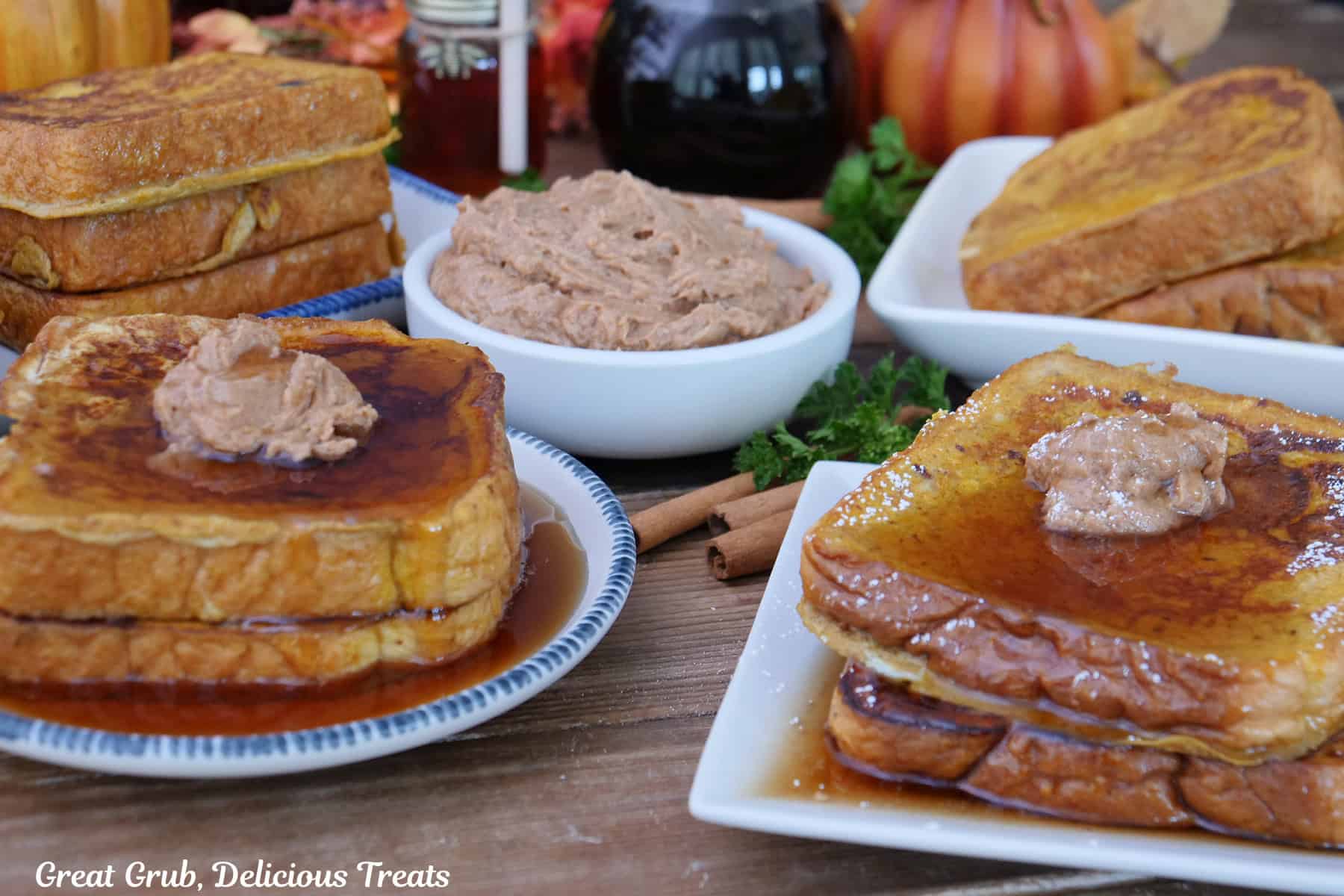 Two plates with two slices of pumpkin spice French toast with cinnamon honey butter and maple syrup on top, with more French toast in the background and a small white bowl filled with cinnamon honey butter.