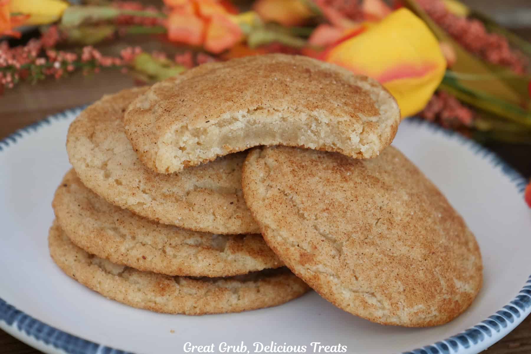 A white plate with blue trim with a stack of five pumpkin spice snickerdoodle cookies on it with a bite out of the cookie on top, and fall foliage in the background.
