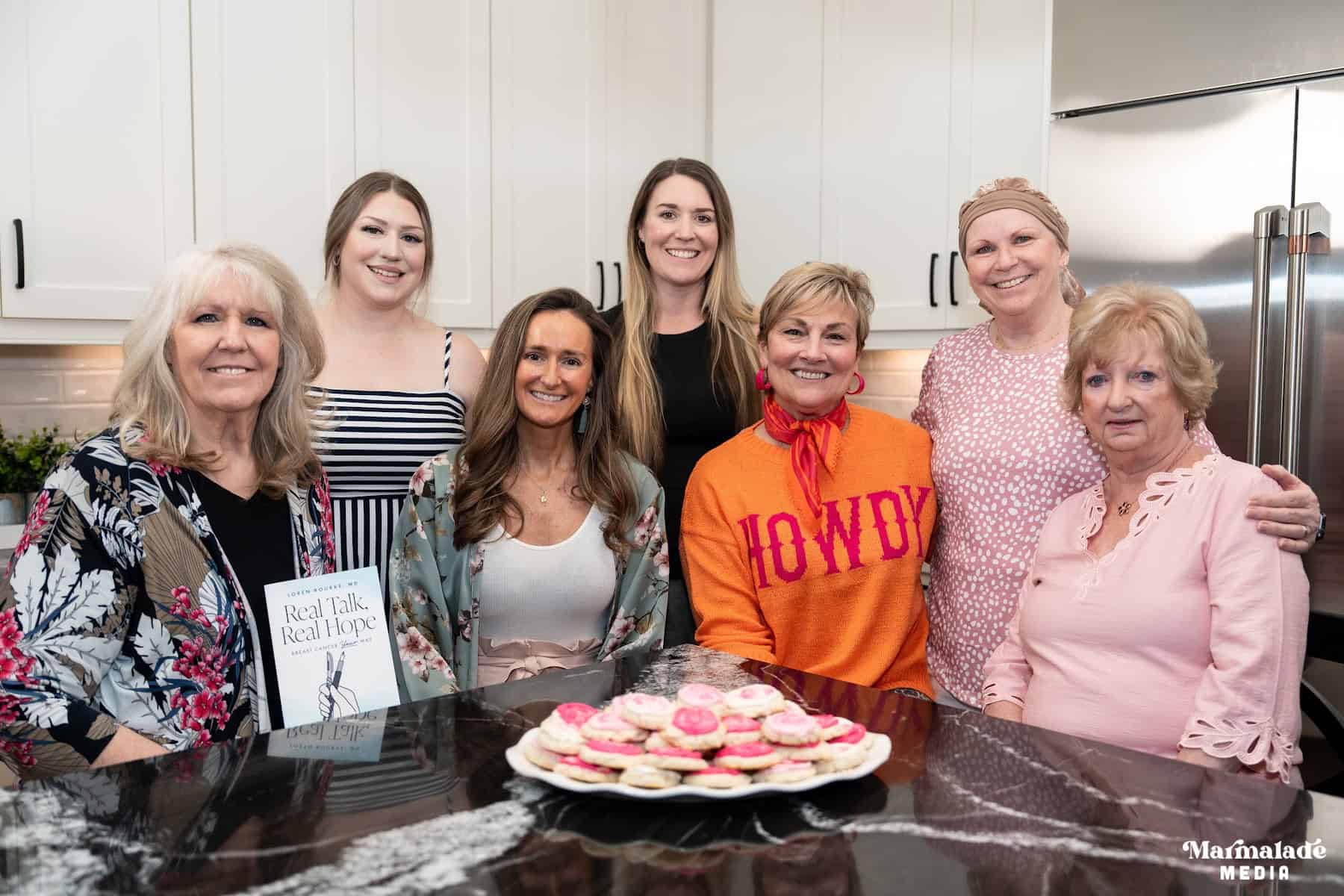 Seven ladies gathered around a kitchen counter with a plate of cookies on the counter, and one holding up a book.