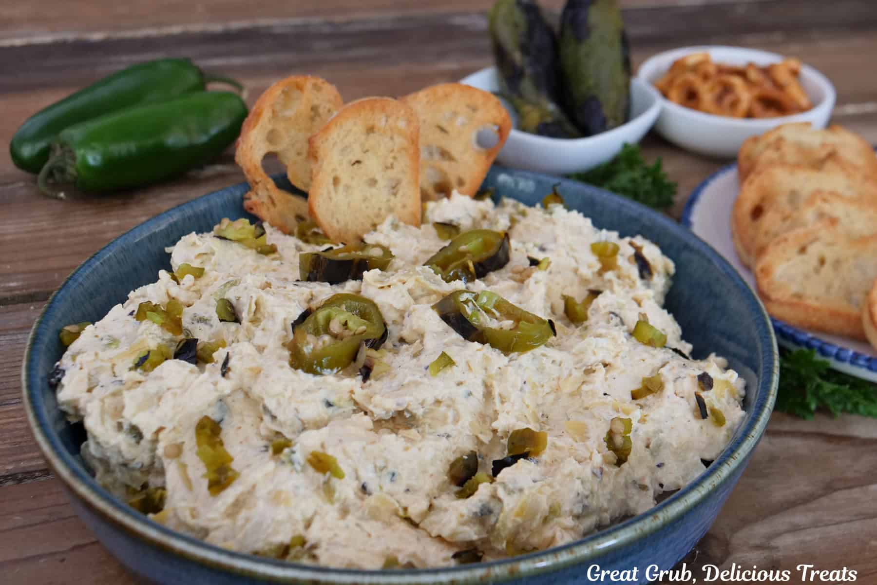Roasted jalapeno artichoke dip in a blue bowl with three crispy bread slices stuck in the dip, and jalapenos and more bread in the background.