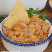 A blue trimmed white bowl filled with street corn dip with a corn tortilla in the dip.