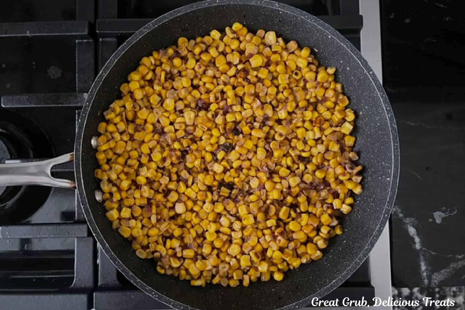 A frying pan on the stovetop with roasted corn and onions in it.