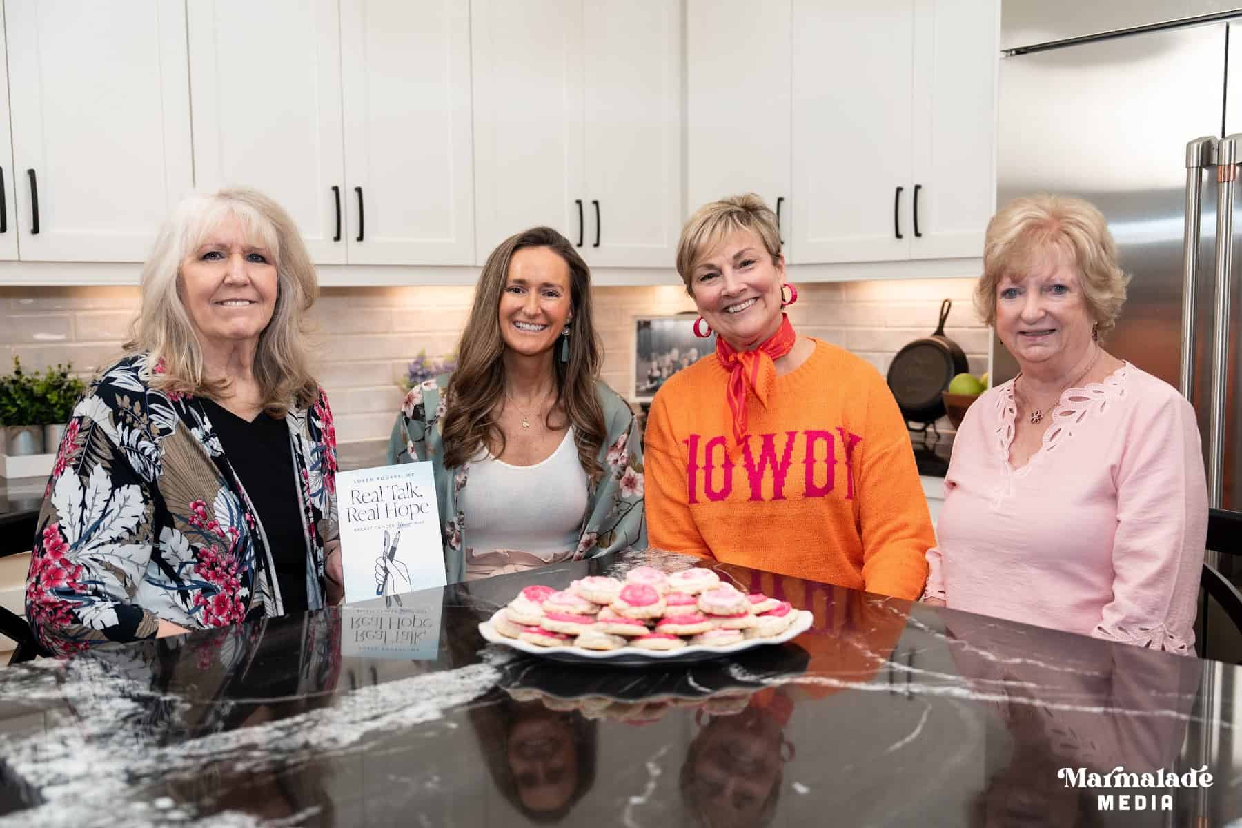 Four women sitting at a kitchen counter with a plate of cookies, and one holding a book.